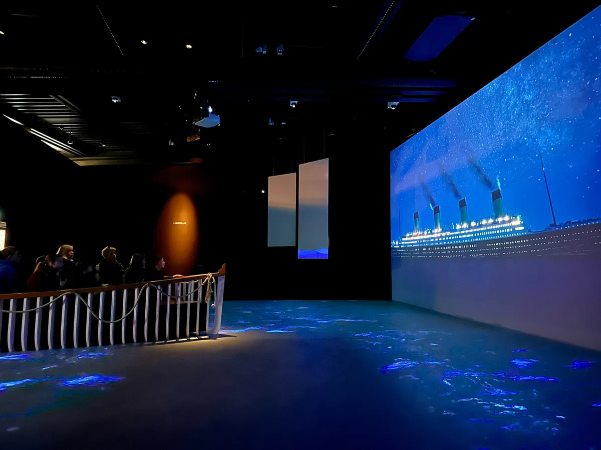 Eine Gruppe von Menschen betrachtet eine beleuchtete Projektion der Titanic in einem dunklen Raum.