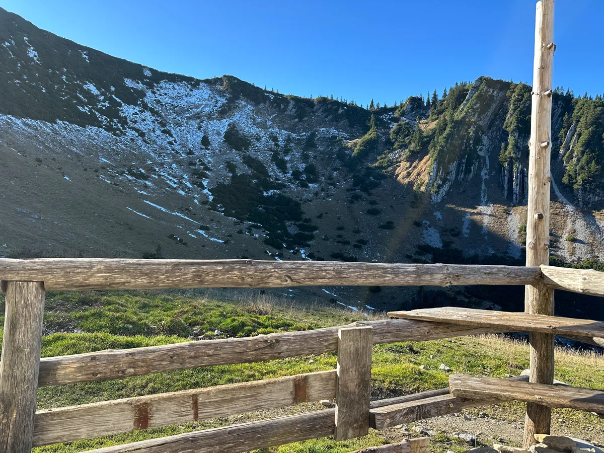 Ein Holzgeländer der Jägerbauernalm mit Blick auf den Jägerkamp, mit vereinzelten Schneeresten unter klarem, blauem Himmel.