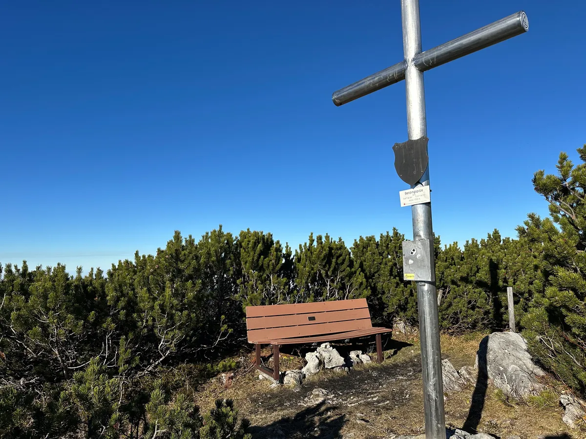 Das Gipfelkreuz der Benzingspitze steht neben einer Holzbank, umgeben von grünen Sträuchern unter einem strahlend blauen Himmel.