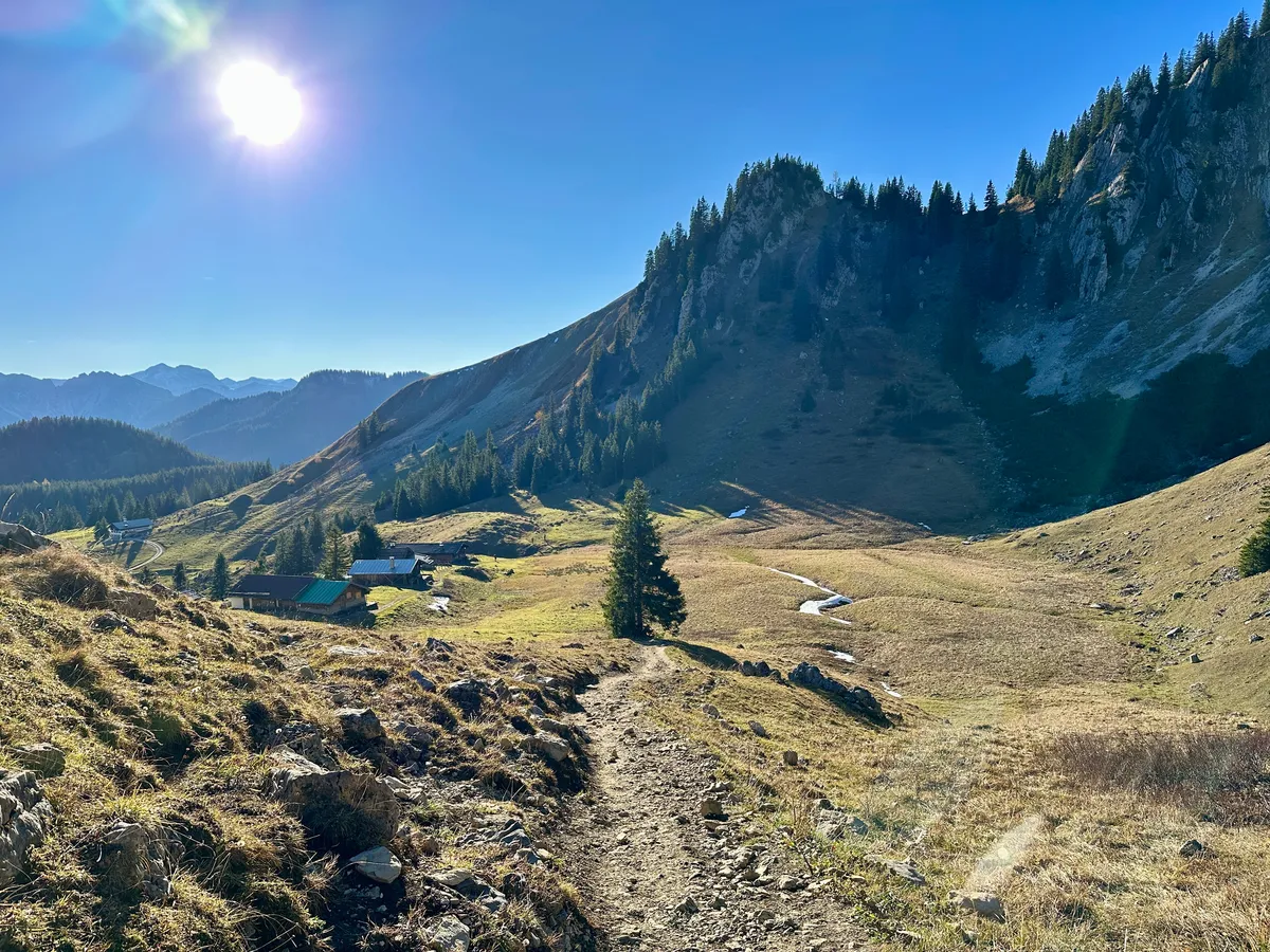 Eine malerische Berglandschaft mit Blick auf das Wilde Fräulein, zeigt einen sonnigen Tag mit einem klaren Weg, der durch ein alpines Tal mit einer Alm führt.