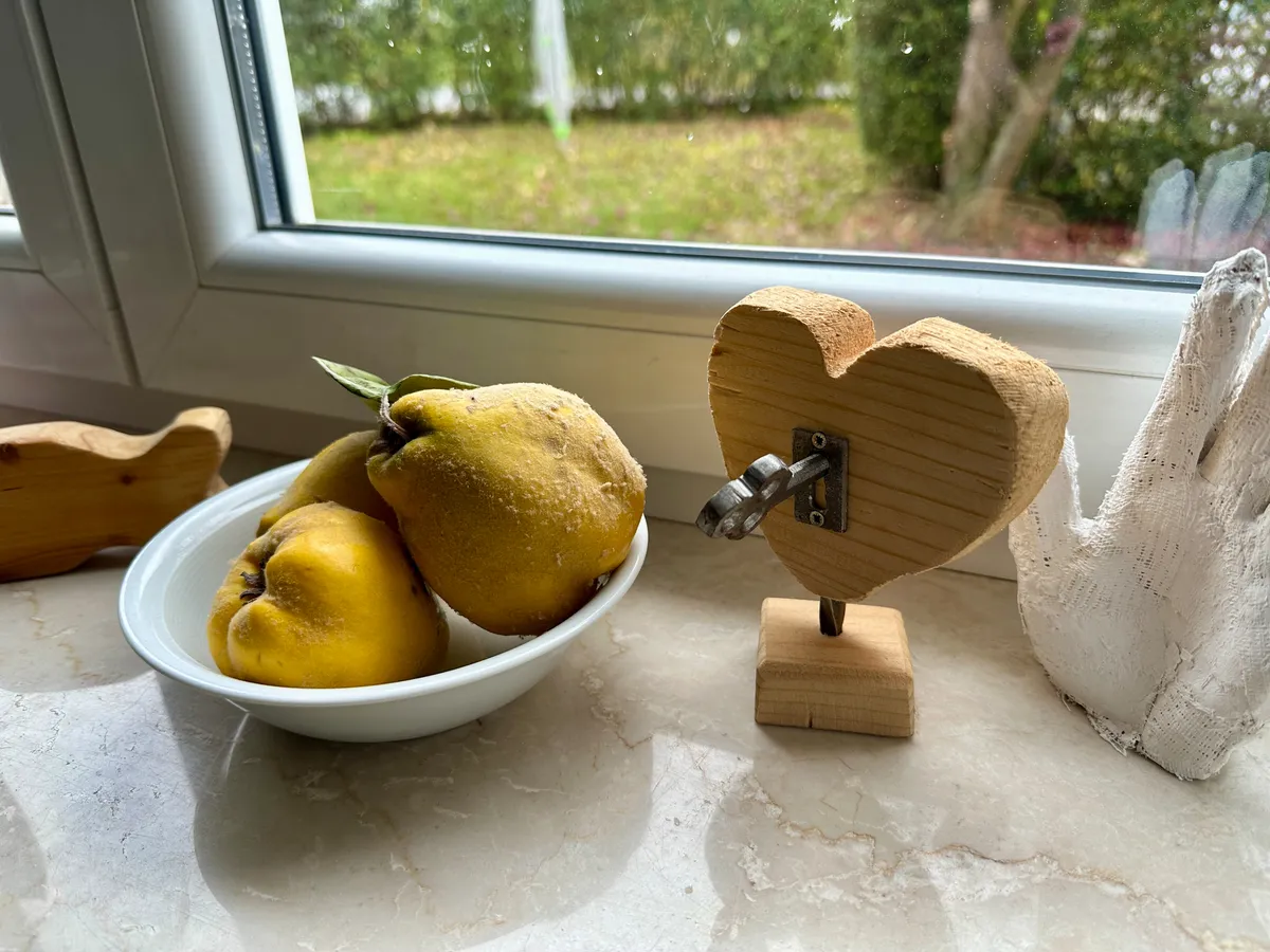 Eine weiße Schale mit drei gelben Quitten steht neben einem herzförmigen Holzdekoration auf einer Fensterbank.