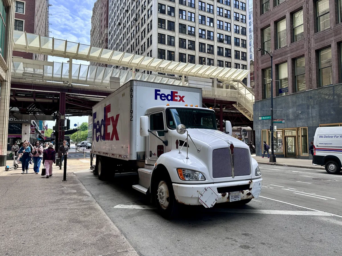Ein weißer FedEx-Lkw steht auf einer städtischen Straße, umgeben von hohen Gebäuden. Fußgänger gehen auf dem Bürgersteig vorbei, während eine Überführung in der Nähe sichtbar ist.