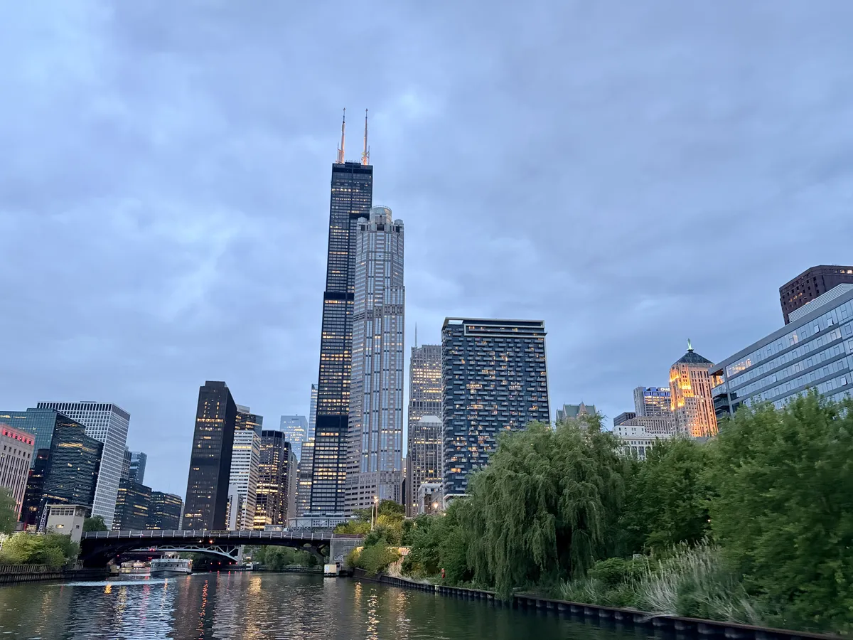 Die Skyline einer Stadt bei Dämmerung, mit hohen Wolkenkratzern, darunter der Willis Tower, der durch seine charakteristischen Spitzen hervorsticht. Im Vordergrund fließt ein ruhiger Fluss, umgeben von grünen Ufervegetationen.