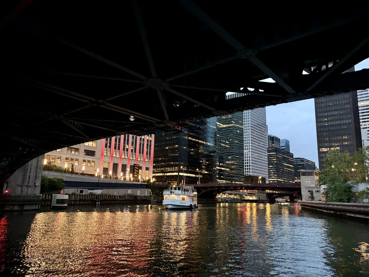 Ein Boot fährt unter einer Brücke hindurch auf einem ruhigen Fluss, der von modernen Wolkenkratzern und beleuchteten Gebäuden gesäumt ist. Die Lichter reflektieren sich im Wasser, während der Himmel in Dämmerlicht getaucht ist.