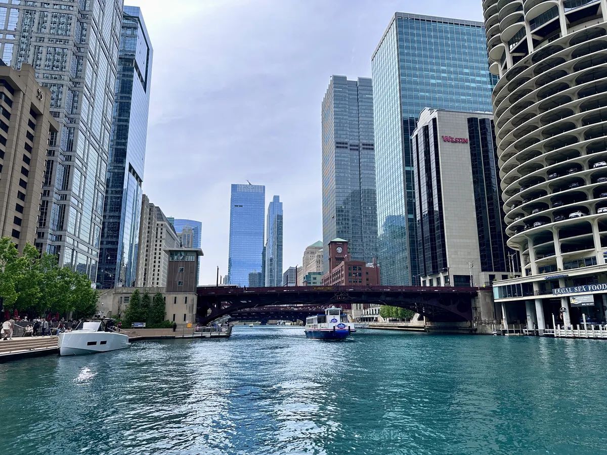 Die Szene zeigt einen Blick auf einen Stadtfluss, der von modernen Wolkenkratzern flankiert wird. Eine Brücke überspannt den Fluss, während ein Boot vorbeifährt und Bäume entlang der Uferpromenade sichtbar sind.
