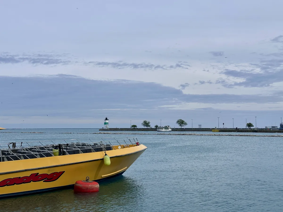 Ein gelbes Boot mit der Aufschrift „Speedog“ liegt im ruhigen Wasser, während im Hintergrund ein Leuchtturm und ein Hafen mit einigen Bäumen und einer Mauer sichtbar sind. Der Himmel ist bewölkt.
