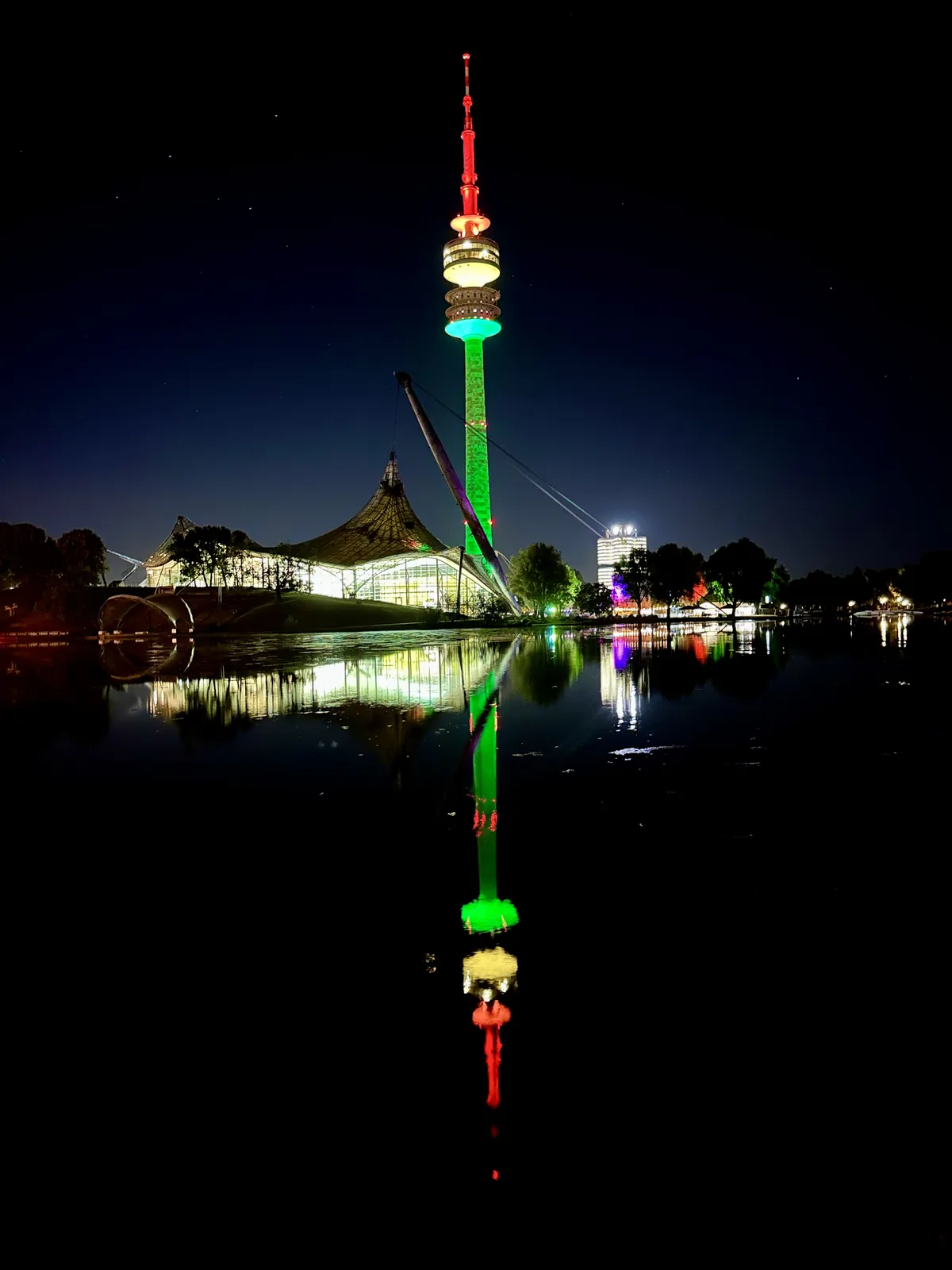 Der beleuchtete Fernsehturm steht bei Nacht am Ufer eines Gewässers. Seine farbigen Lichter reflektieren sich im ruhigen Wasser, während ein festivales Zelt im Hintergrund sichtbar ist.