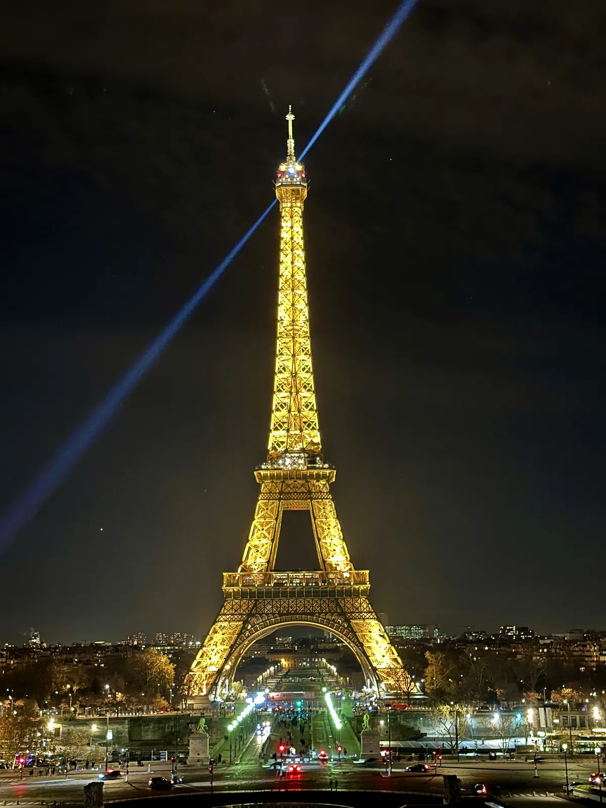 Der Eiffelturm in Paris erstrahlt nachts in goldenem Licht mit einem leuchtenden Suchscheinwerfer.