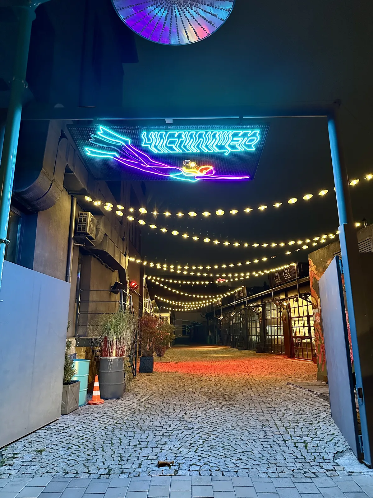 Ein gepflasterter Durchgang ist nachts von bunten Lichterketten und einem neonblauen Schild beleuchtet.