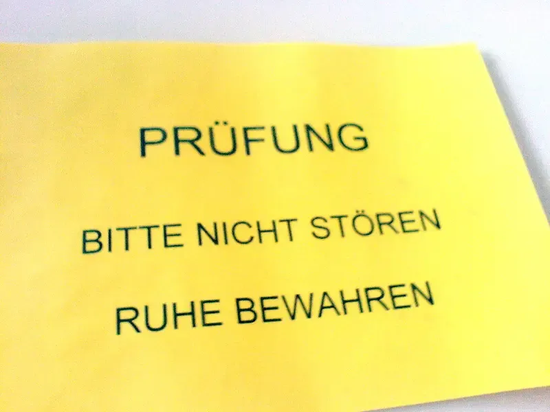 Ein gelbes Schild mit schwarzer Schrift, das die Worte "PRÜFUNG", "BITTE NICHT STÖREN" und "RUHE BEWAHREN" enthält.