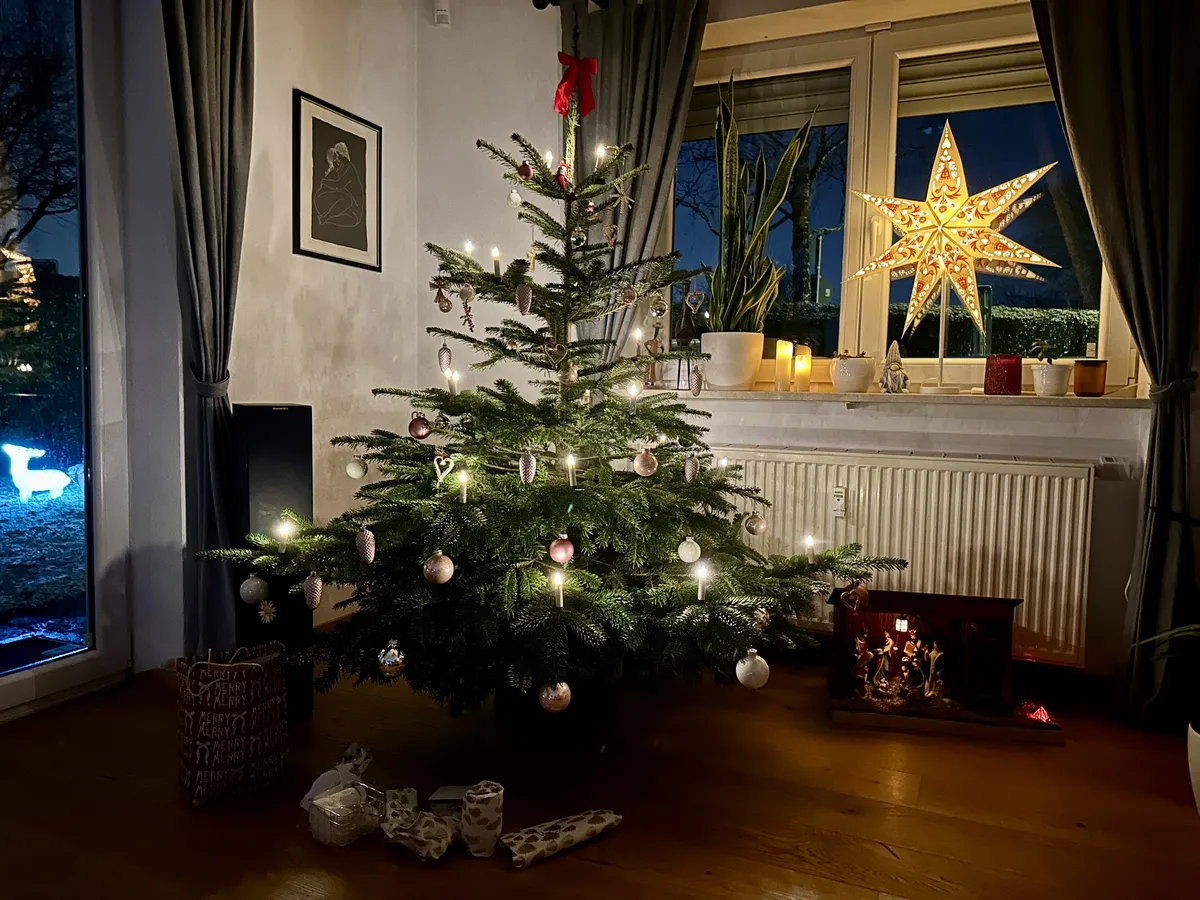 Ein festlich geschmückter Weihnachtsbaum mit warmweißen Kerzen Lichtern und rosa-silbernen Kugeln steht in einem gemütlichen Wohnzimmer. Im Hintergrund leuchtet ein großer Papierstern am Fenster neben einer Krippe und Zimmerpflanzen.