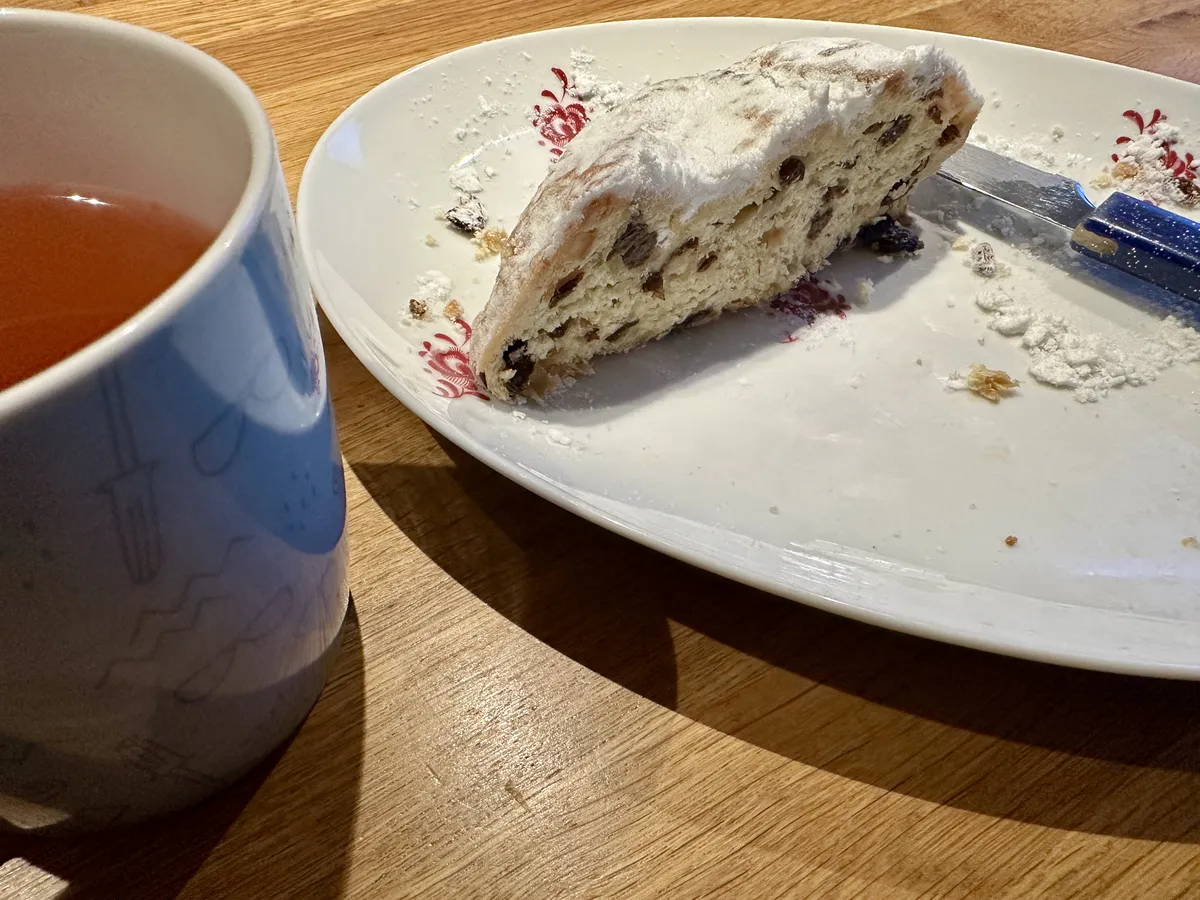 Auf einem Holztisch liegt ein Stück Stollen, das mit Puderzucker bestäubt ist, neben einer Tasse Tee. Der Stollen enthält Trockenfrüchte und Nüsse und ist auf einem dekorativen Teller platziert.