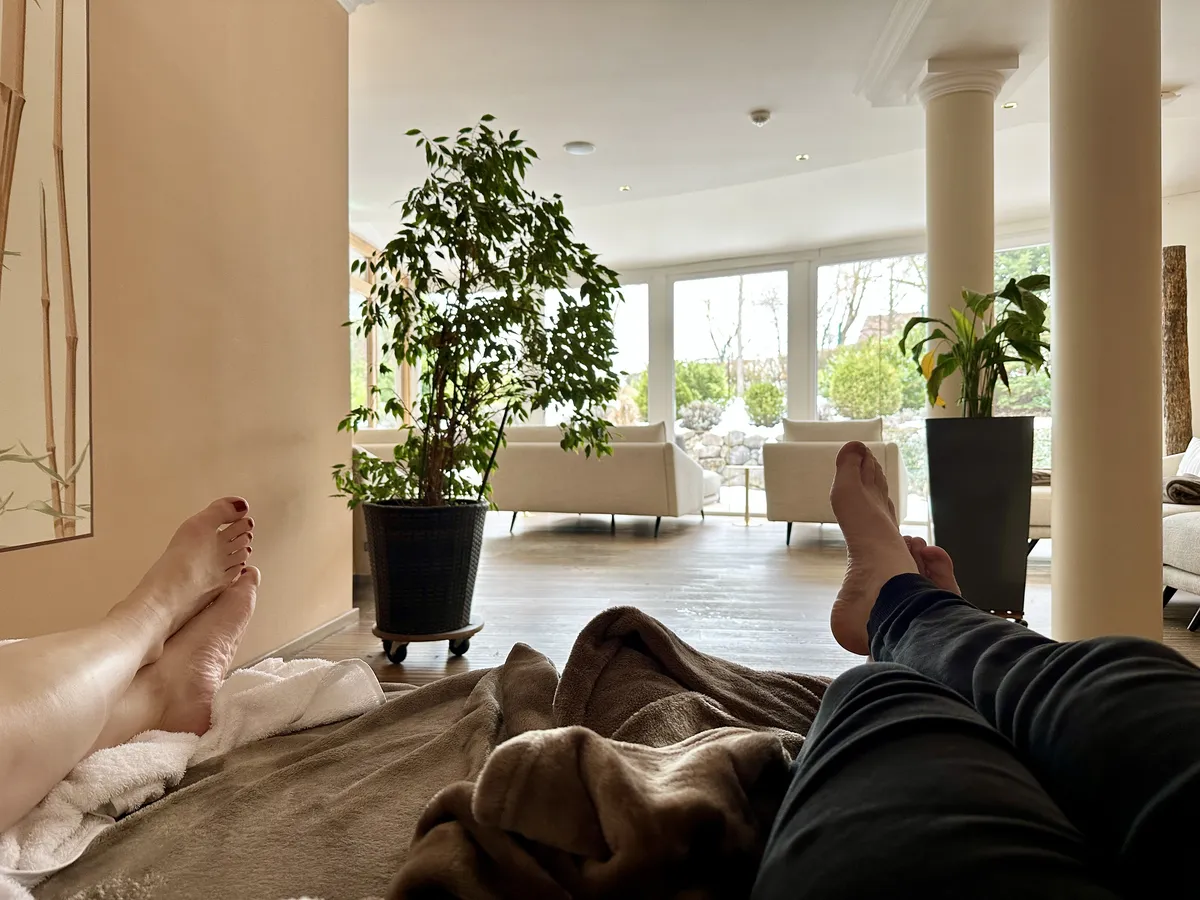 Zwei Menschen liegen entspannt auf einer Decke. Im Hintergrund ist ein heller Raum mit großen Fenstern, Pflanzen und einer gemütlichen Sitzgelegenheit zu sehen.