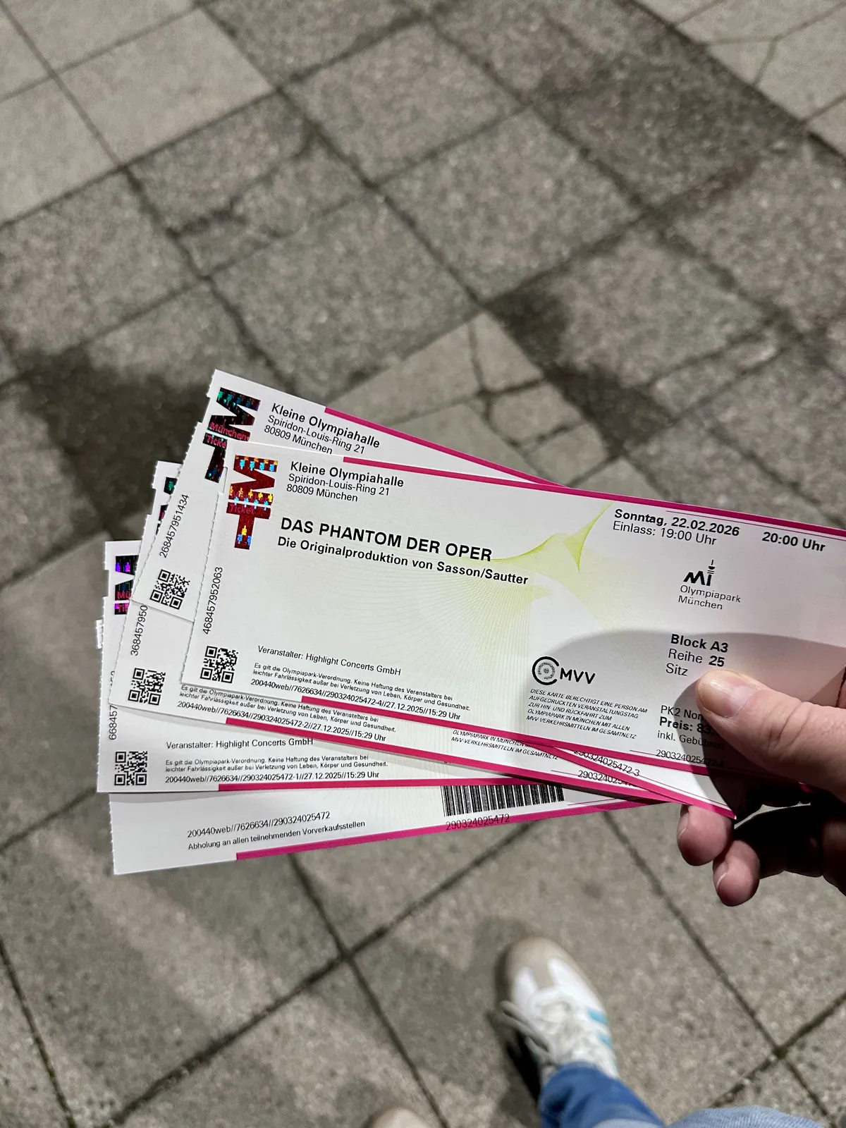 Mehrere Tickets für das Musical "Das Phantom der Oper" sind in einer Hand zu sehen, mit einem klaren Fokus auf das Event-Datum und die Veranstaltungsdetails. Im Hintergrund sind graue Pflastersteine sichtbar.