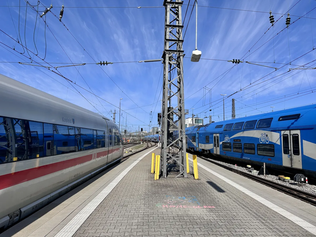 Zwei Züge stehen an einem Bahngleis, einer auf der linken Seite in weiß-roter Farbe und der andere auf der rechten Seite in Blau. Der Himmel ist klar mit einigen Wolken und elektrische Masten sind im Hintergrund sichtbar.