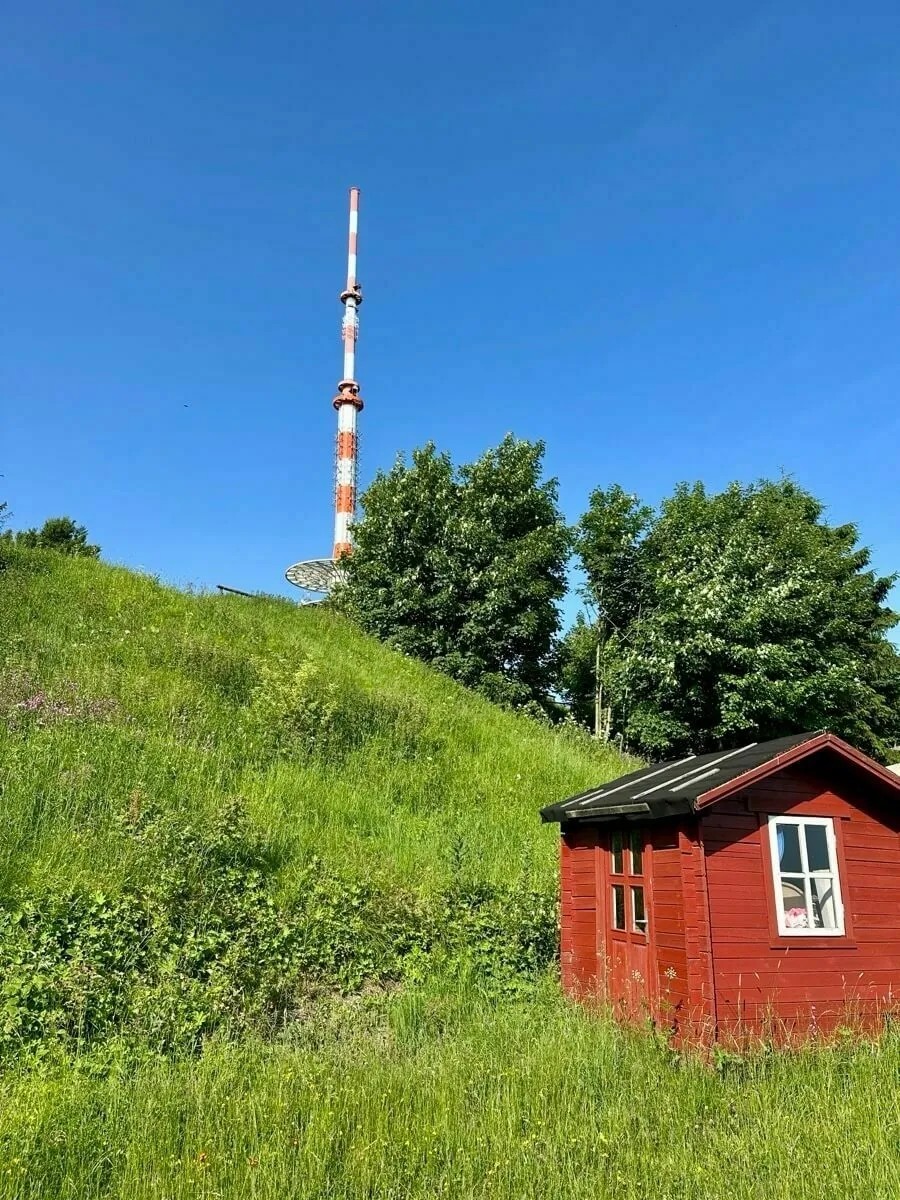 Ein kleines rotes Häuschen steht auf einer grünen Wiese neben einem Hügel mit einem großen Funkmast und Bäumen im Hintergrund.
