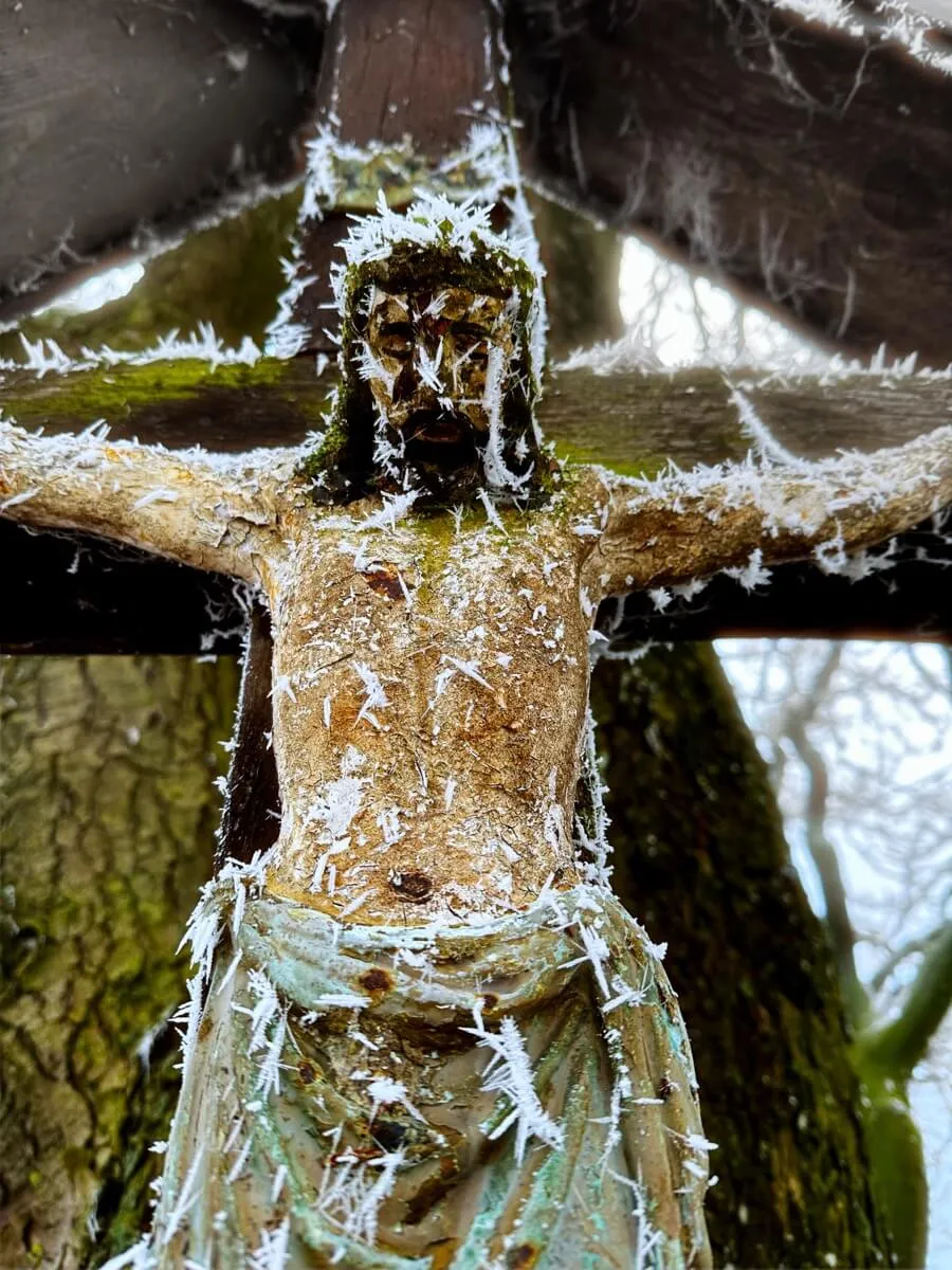 Eine frostbedeckte Christusfigur am Kreuz, umgeben von weißen Eiskristallen und Moos, steht im Vordergrund. Im Hintergrund sind verschwommene Bäume und eine winterliche Landschaft sichtbar.