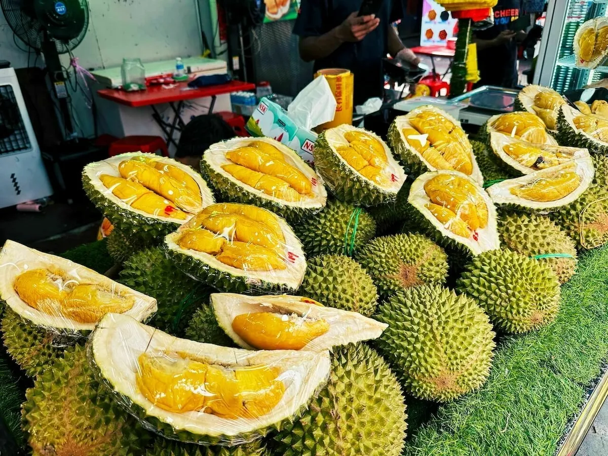 Ein Marktstand in Kuala Lumpur mit einem großen Stapel von Durian-Früchten. Viele der stacheligen Früchte sind aufgeschnitten, um das leuchtend gelbe Fruchtfleisch zu zeigen, das einzeln in Plastikfolie verpackt ist.
