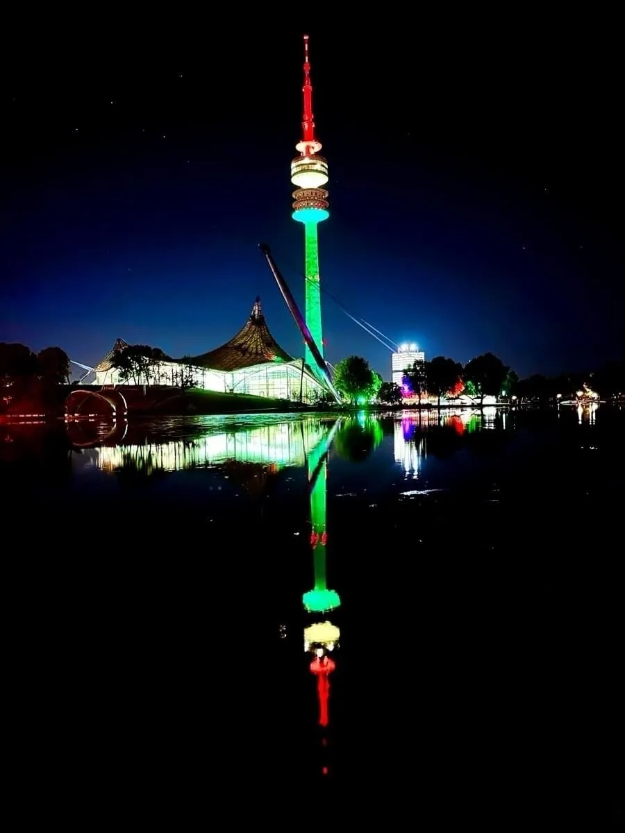 Der Münchner Olympiaturm mit farbiger Beleuchtung spiegelt sich im Olympiasee in der Nacht wieder.
