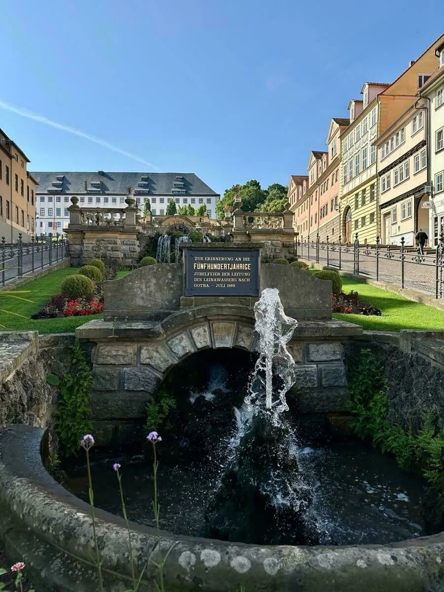 Ein Springbrunnen mit einer Inschrift im Vordergrund befindet sich vor einer Reihe farbenfroher Gebäude und dem historischen Rathaus von Gotha unter einem blauen Himmel.