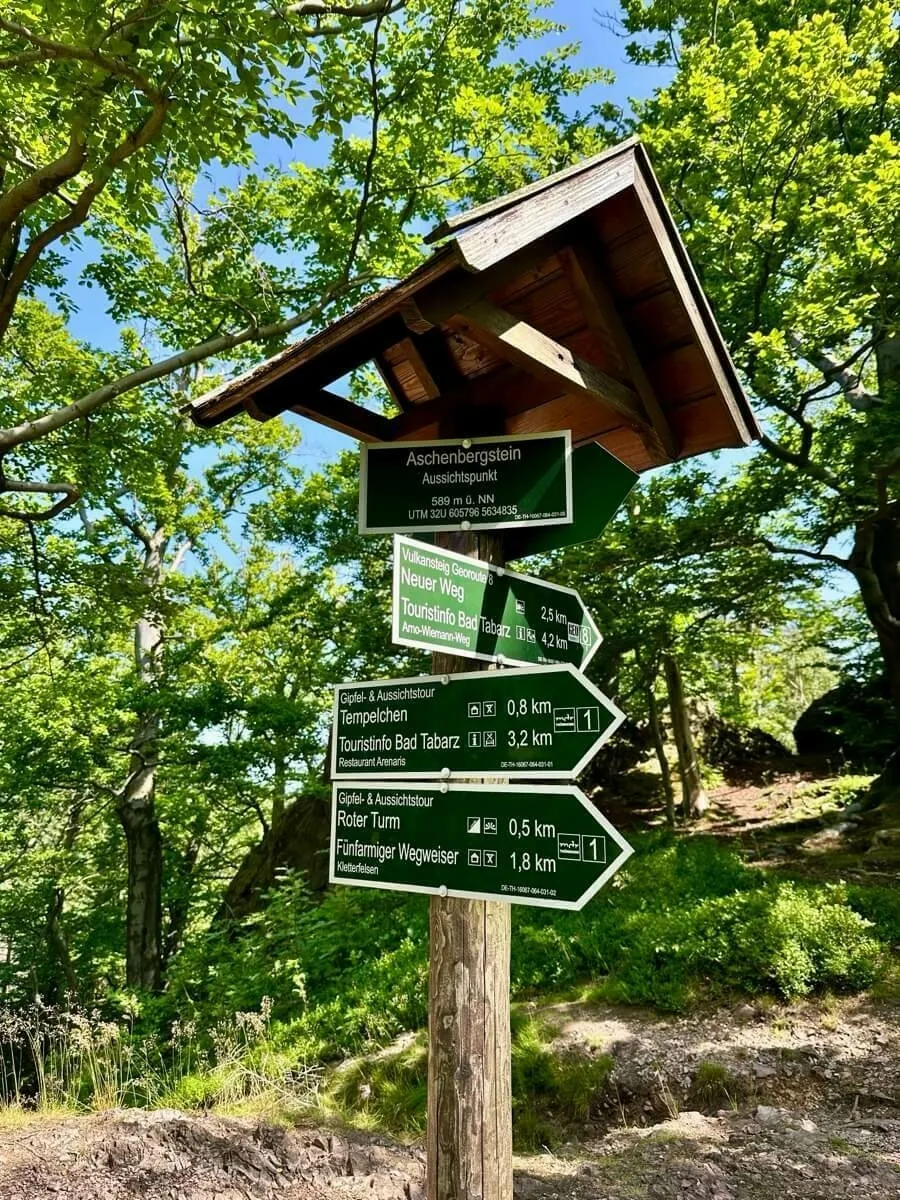 Ein hölzernes Wegweiser zeigt mehrere Wanderwege und Entfernungen im Waldgebiet an.