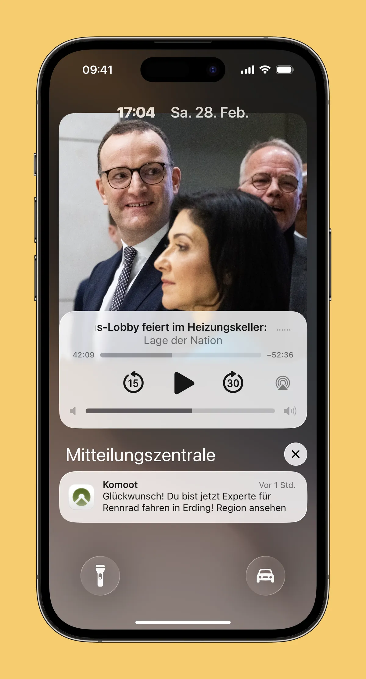 Auf dem Bildschirm eines Smartphones ist eine Audio-Wiedergabe angezeigt, die Titel und Fortschrittsbalken des Podcasts "Lage der Nation" zeigt. Im Hintergrund ist eine Gruppe von Menschen zu sehen, darunter ein Mann mit Brille, der in einem Anzug steht. Unterhalb der Wiedergabe ist eine Benachrichtigung von Komoot, die eine Glückwunschmeldung enthält.