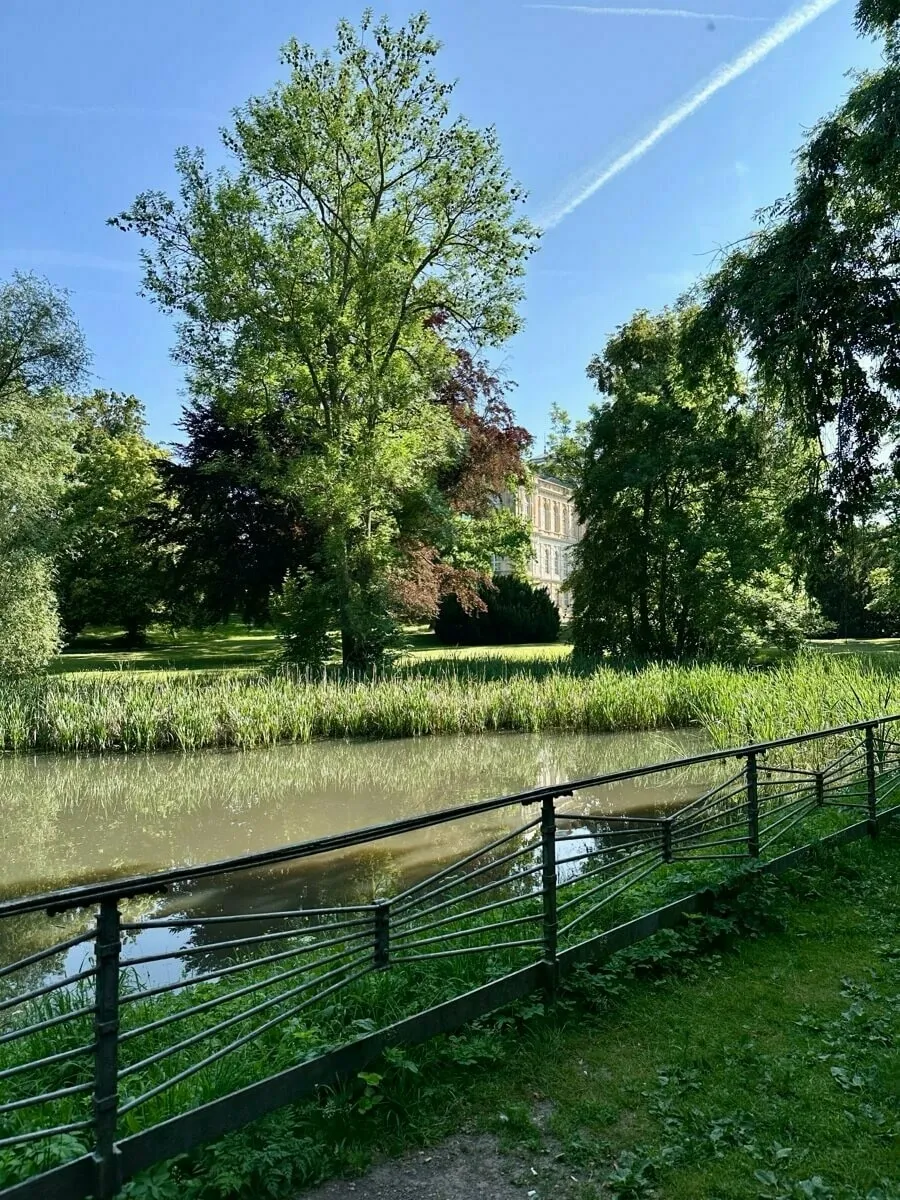 Ein ruhiger Teich mit einem Zaun im Vordergrund und dem herzoglichen Museum im Hintergrund, umgeben von üppigen, grünen Bäumen.