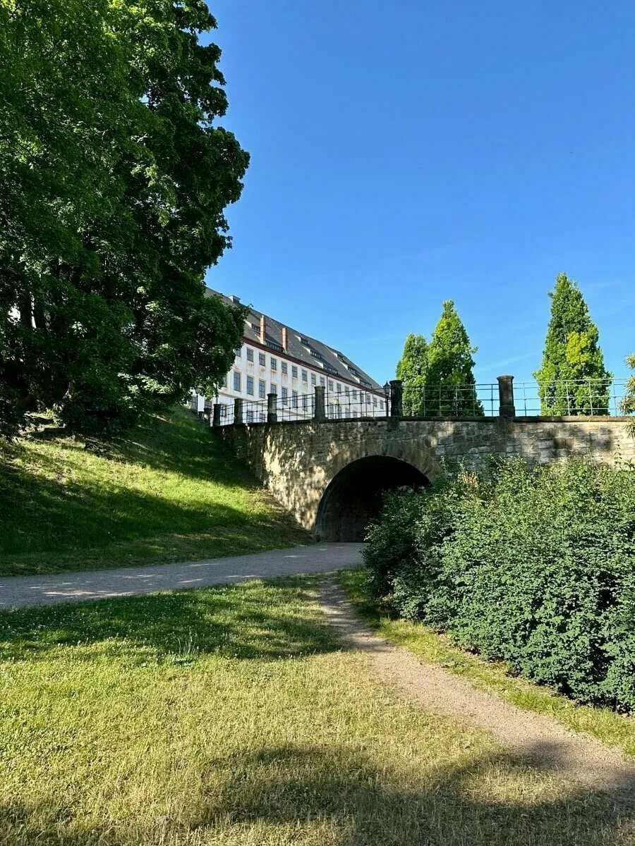 Ein Sandweg führt zu einer Brücke, die an einem sonnigen Tag von Bäumen umgeben ist, mit dem Schloss Friedenstein im Hintergrund.