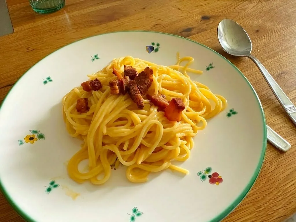 Ein Teller mit Spaghetti Carbonara steht auf einem Holztisch mit einem Löffel daneben.