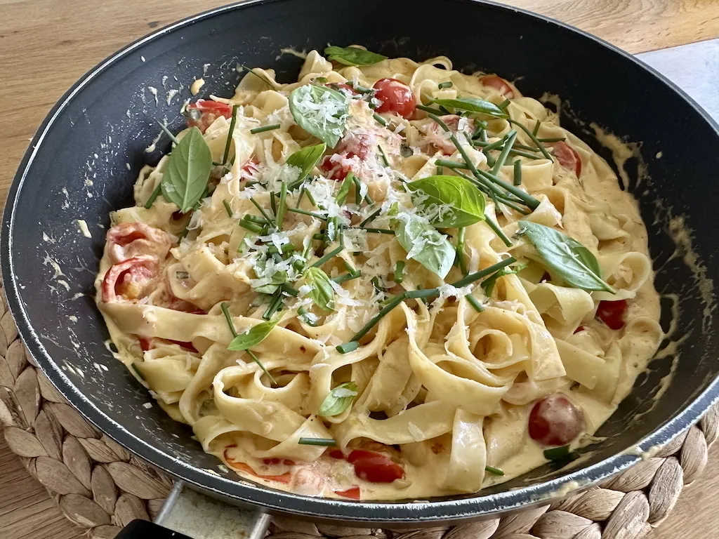 Eine Pfanne mit cremigen Nudeln, Kirschtomaten und frischen Basilikumblättern, bestreut mit geriebenem Käse.