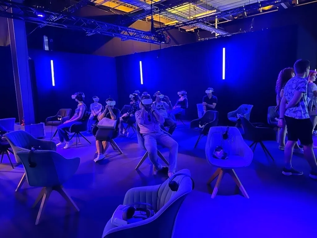 Eine Gruppe von Menschen sitzt in bequemen Stühlen in einem dunkelblauen Raum mit Neonröhren und trägt VR-Brillen für eine immersive Erfahrung in der Titanic-Ausstellung.