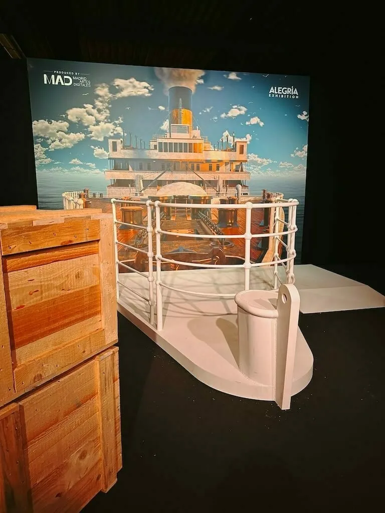 Ein Ausstellungsstück, das einen Teil des Decks der Titanic mit weißem Geländer und einer Rettungsboothalterung vor einer großen Fototapete des Schiffes darstellt. Daneben stehen gestapelte Holzkisten.