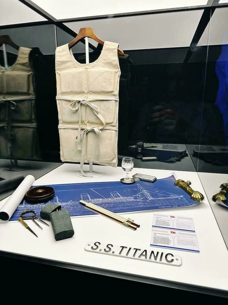 Eine Vitrine mit nachgebildeten Artefakten der Titanic, einschließlich einer historischen Schwimmweste, einer blauen Schiffsbauzeichnung und Besteck. Ein kleines Schild trägt die Aufschrift "S.S. TITANIC".