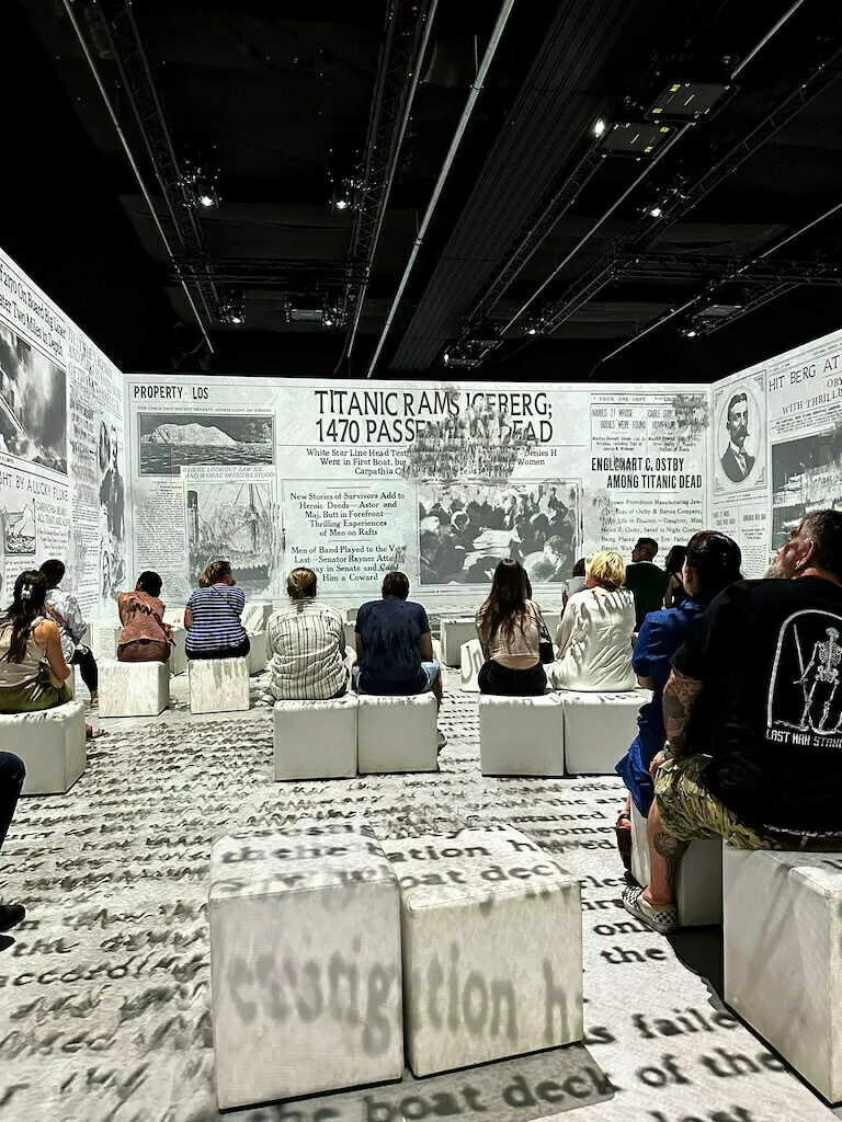 Besucher sitzen in einer Reihe auf weißen Hockern vor einer großen Wand, die mit reproduzierten Zeitungsausschnitten über den Untergang der Titanic tapeziert ist, mit der Schlagzeile: "TITANIC RAMS ICEBERG; 1470 PASSENGER DEAD".