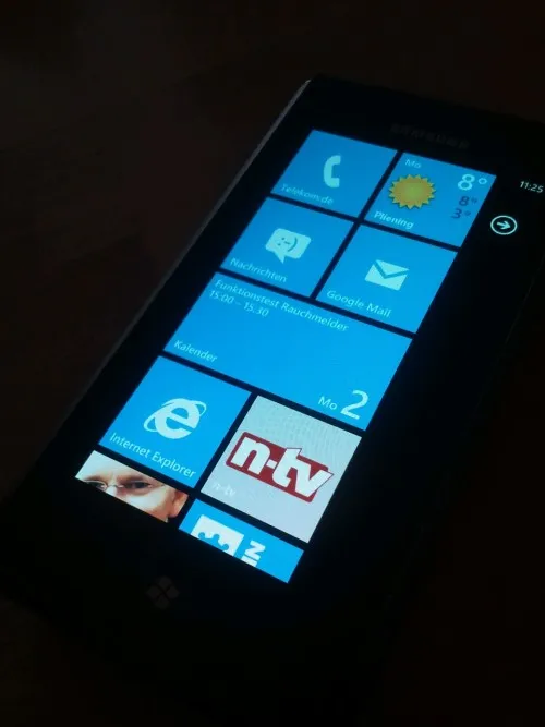 Ein schwarzes Smartphone mit einem blauen Startbildschirm, der Apps wie Telefon, Nachrichten, Google Mail und Internet Explorer anzeigt. Die Uhrzeit ist 11:25 und es gibt ein Wetterwidget mit einer Sonnensymbolik.