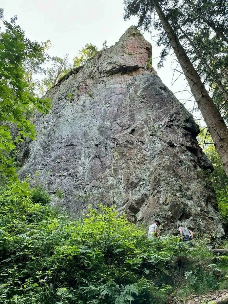 Ein großer Felsen erhebt sich in einem bewaldeten Gebiet, während zwei Personen davor stehen.