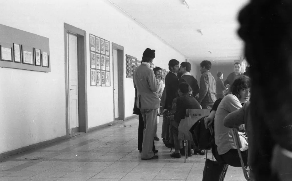 A 1974 black and white photograph of a long institutional corridor during visiting hours at a boys reformatory in Aszód, Hungary. Teenage boys sit on chairs facing visitors, and in the center two boys lean toward each other and share a brief kiss.