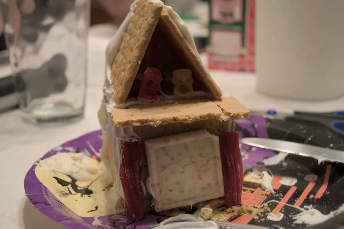 A humble gingerbread house. A Ghirardelli chocolate square acts as the front door. Two gummy bears stand on the second floor underneath the roof.