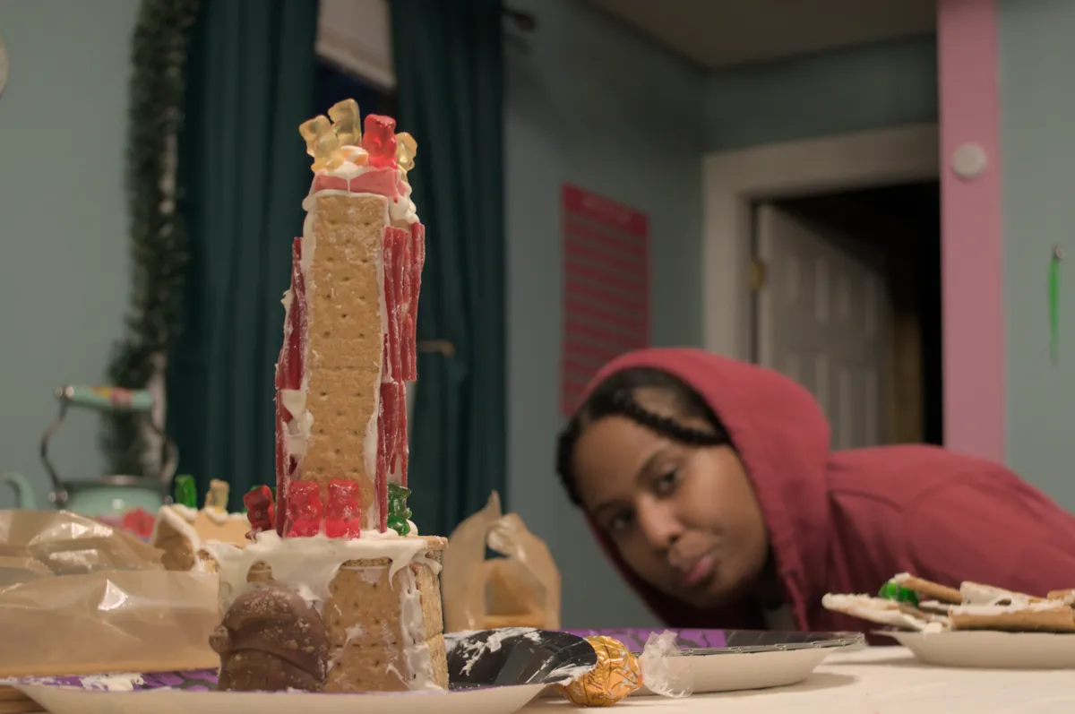 A tower made of graham crackers, white icing, licorice, and gummy bears sits atop a paper plate. It has a larger base with the head of a chocolate Santa on the side. Atop that base is a longer and thinner portion lined with licorice. Gummy bears sit precariously on the top. Amaya photobombs in the background.