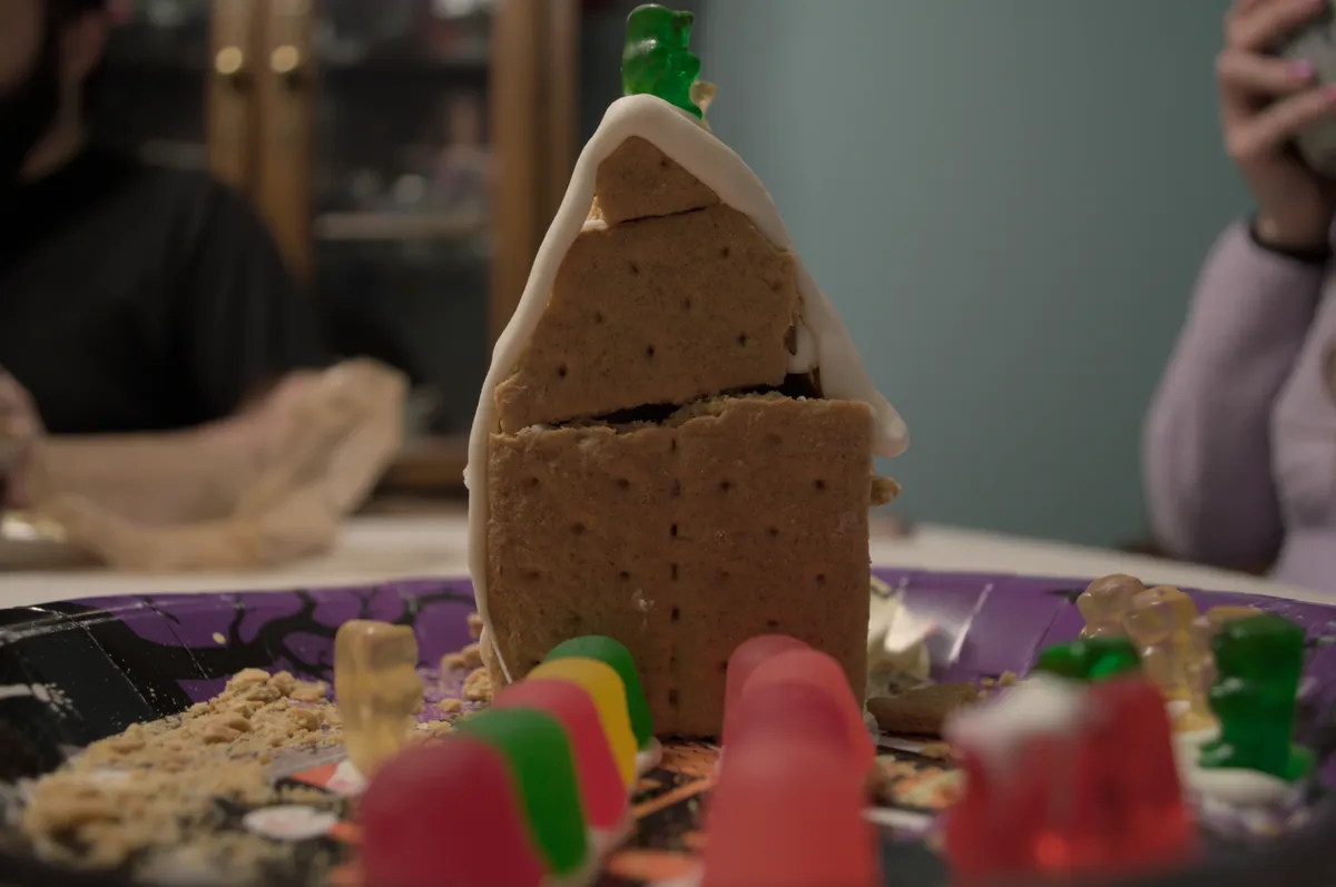 Lines of Dots (the candy) lead up to the graham cracker house. Three gummy bears sit atop it, while several onlookers are off to the side.