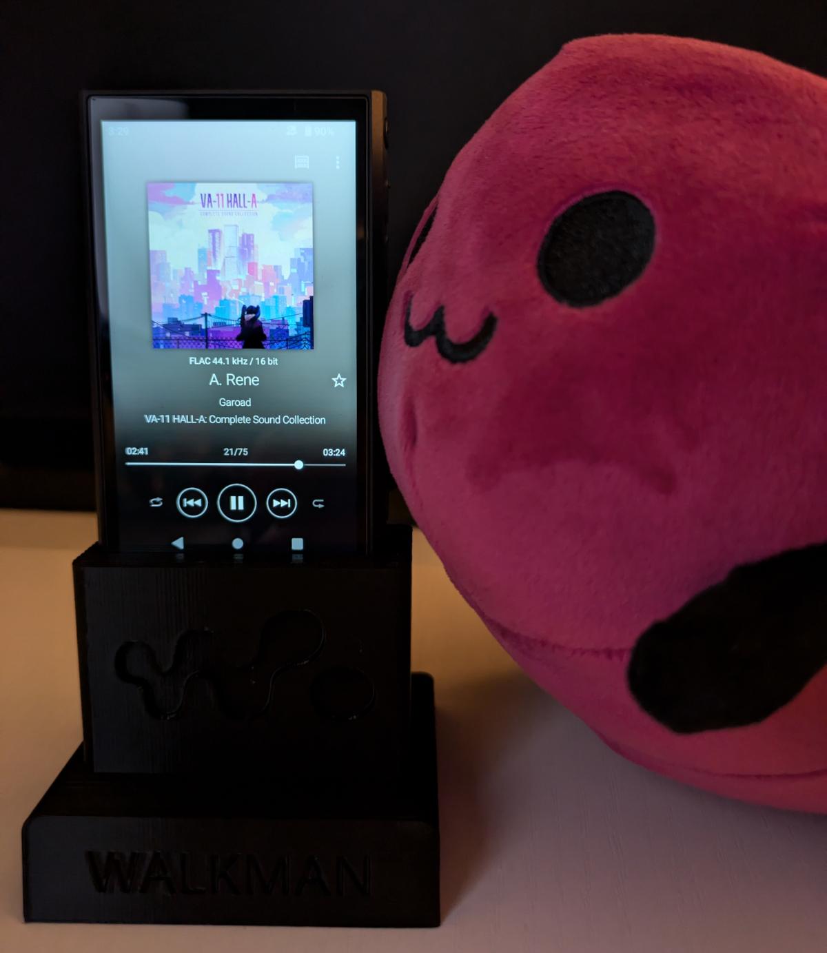 eggbug plushie sitting up against a digital Sony Walkman resting inside a custom 3D printed dock. A. Rene from the Valhalla Cyberpunk Bartender soundtrack is playing on the device.
