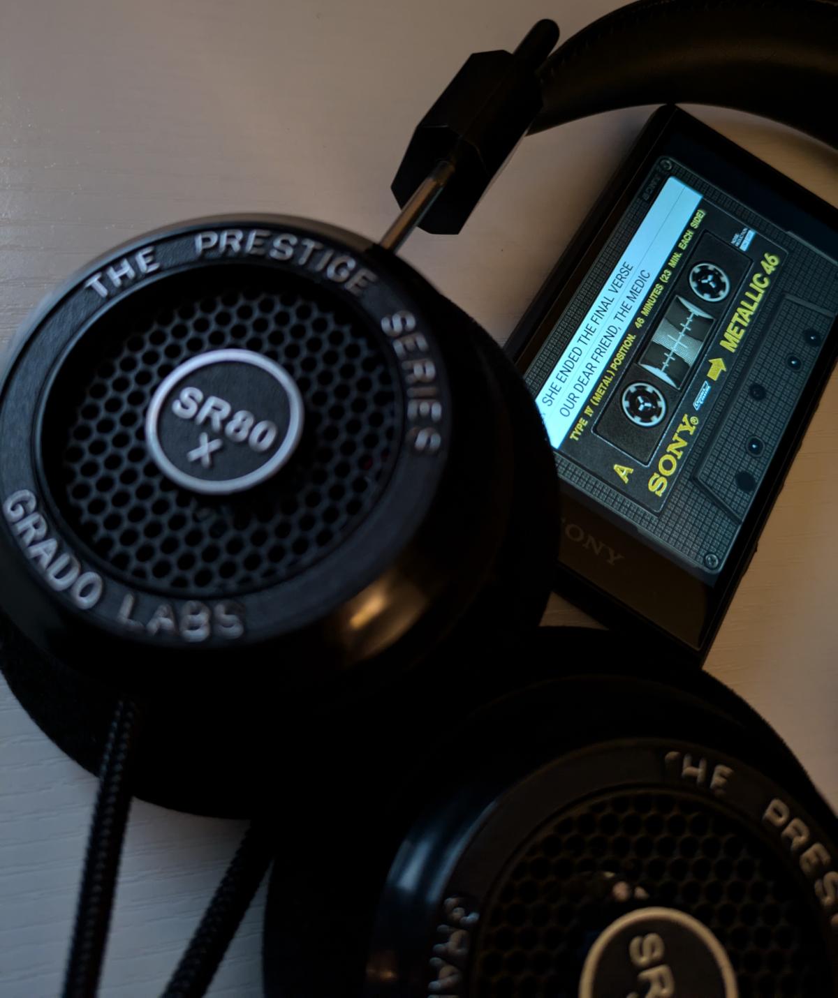 Froey's walkman sat in-between a pair of Grado SR-80x headphones. The song She Ended The Final Verse by Our Dear Friend, The Medic. Is playing in a cassette view.