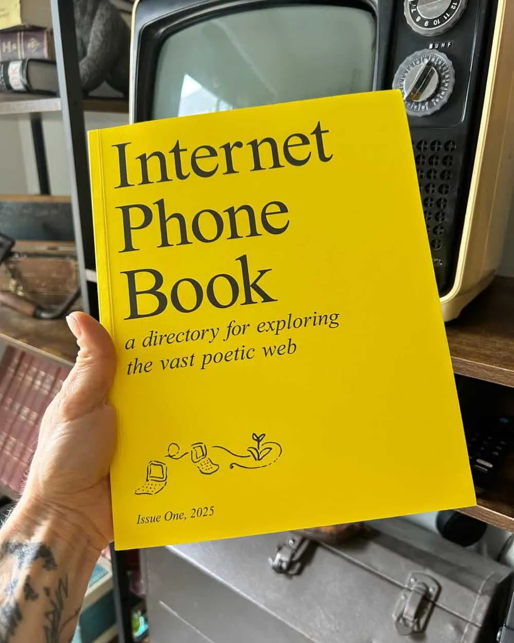 My hand holding up The Internet Phone Book: a directory for exploring the vast poetic web. Issue One, 2025. My black and white Sampo TV is partially visible in the background.