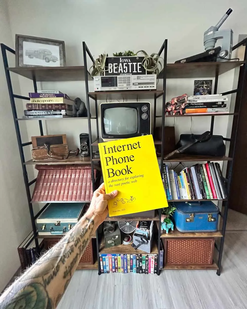 My hand holding up The Internet Phone Book in front my shelving display that holds my 1984 black and white Sampo TV, rows of books, vintage cases, my father's first medical bag, a microscope, and other curiosities that The Internet Phone Book shall now live amongst.
