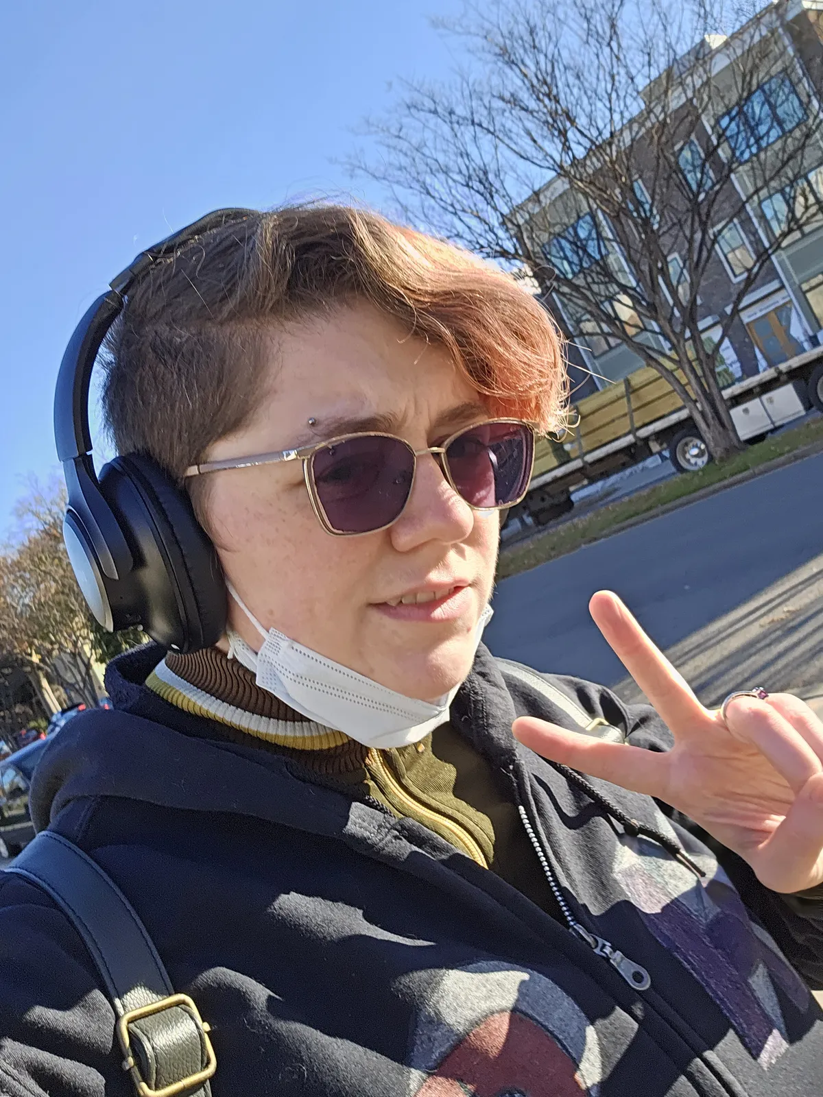 A selfie of a really cool person wearing headphones, subtly smiling and flashing the peace sign. Their hair is cut short, partially bleached and dyed red. Their eyes are obscured by dark transition glasses. Photo taken Jan. 2025