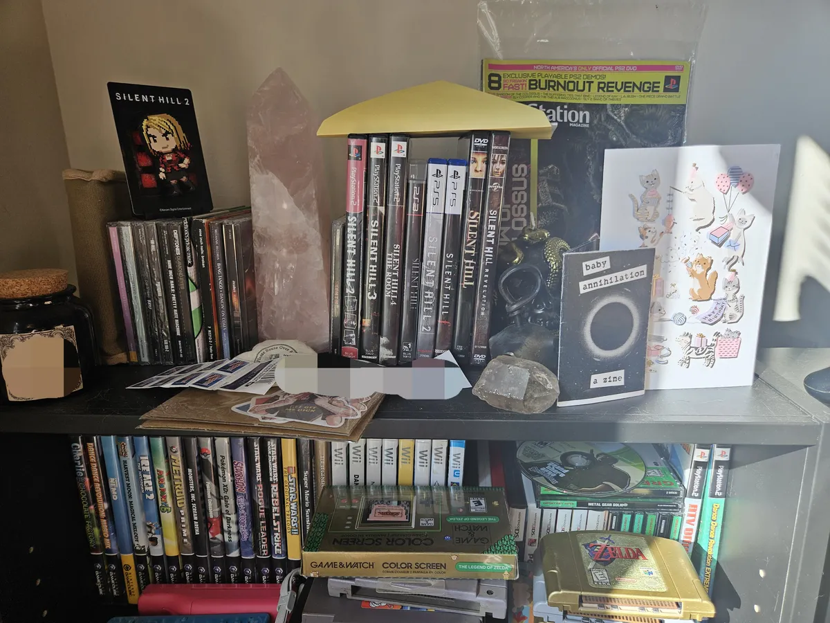 A collection of some Silent Hill videogames and movies that also features music CDs, and older Nintendo games. There's a Playstation magazine that can be spotted in the background, and a small cartoon magnet of Maria from Silent Hill 2 resting on top of the CDs.