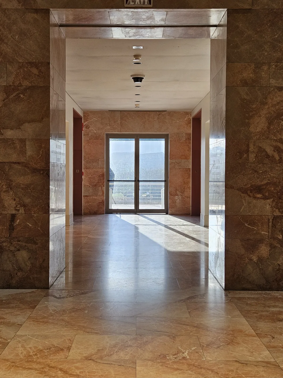 A centered photograph of the entrance to a hallway, accentuated by a doorway to the outside. Light shines through, casting long shadows and reflecting off of pink marble walls and floor.