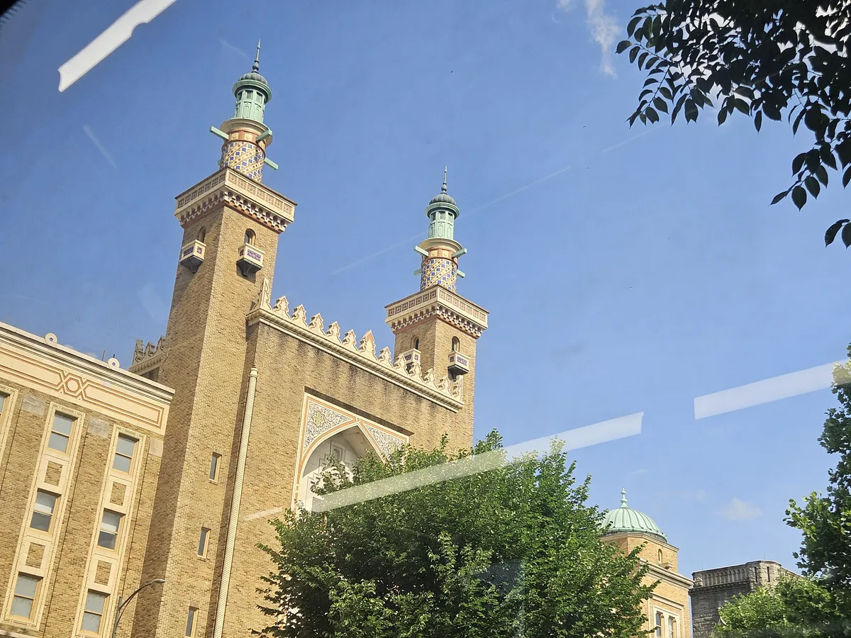 Photograph of the top spires from a notable landmark building. The illusion of an outdoor photo is broken by the reflections of a bus window. Trees frame the right-side of the photo and the sky is cloudless.