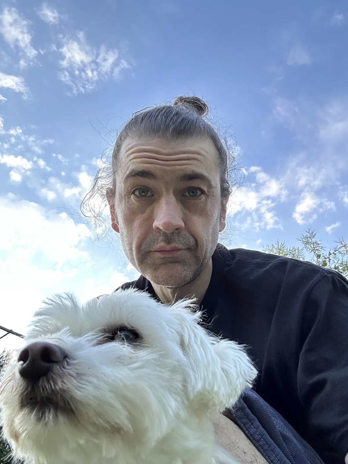 Some middle aged white dude with hair that has been growing for 1983 days. But it’s in a bun. And somehow still manages to be messy. But also there’s a cute white dog in the foreground which should be drawing all your attention.