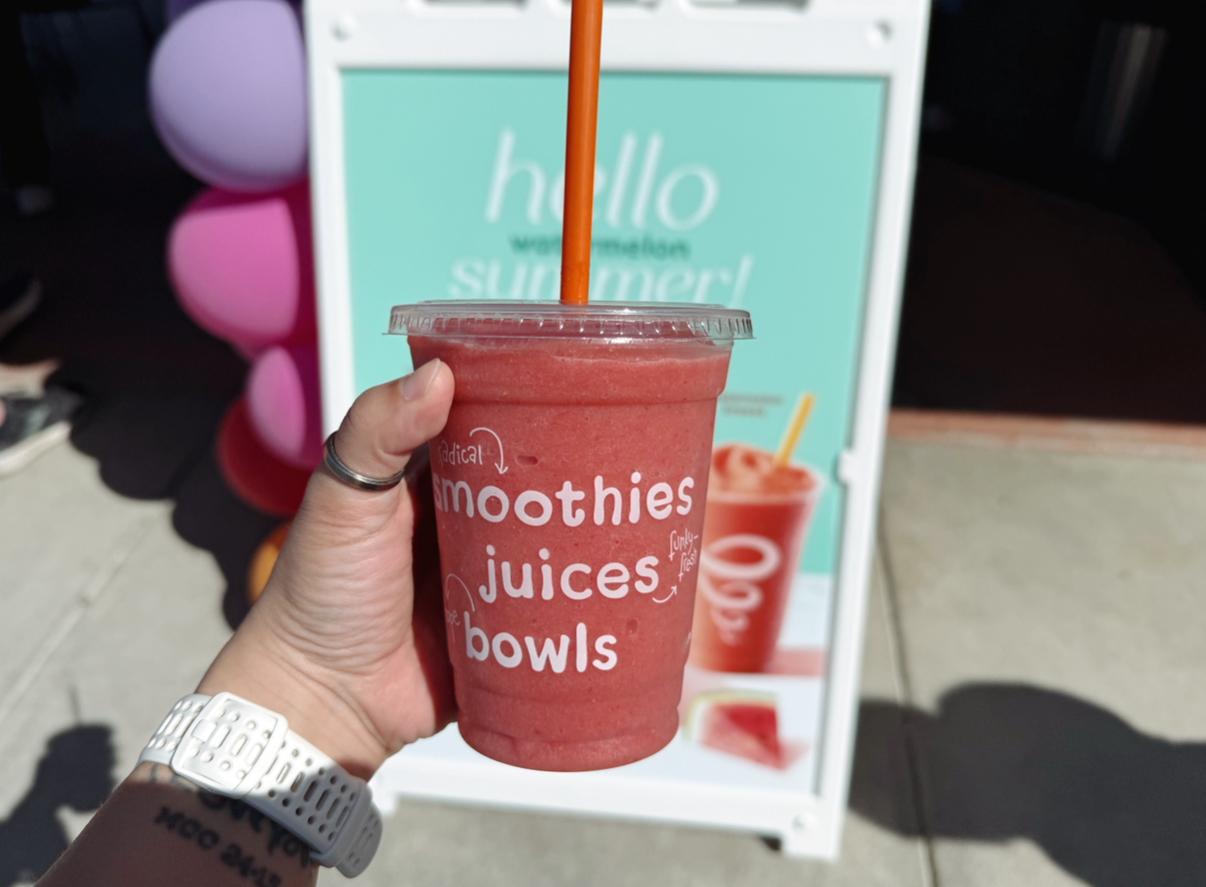 A hand holds a smoothie cup with a red drink and orange straw.